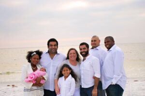 A photograph of a group of people smiling, wearing white shirts standing together next to the beach. A lady is holding a baby in a pink blanket.